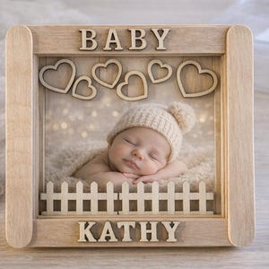 Peut inclure: Un cadre photo en bois avec le mot "BABY" en haut et "KATHY" en bas. Le cadre comporte une petite clôture blanche et des cœurs en bois. À l'intérieur du cadre se trouve un bébé endormi portant un bonnet en tricot.