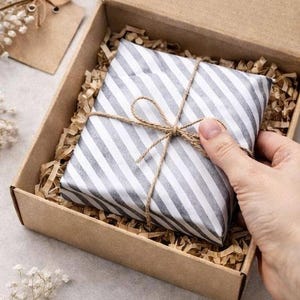 May include: A square gift package wrapped in white and grey striped paper, tied with thin brown twine in a bow. The package rests inside a brown cardboard box filled with shredded paper filler, providing a protective cushion for the item inside.