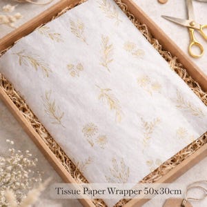 May include: A white tissue paper wrapper with gold botanical designs, measuring 50x30cm, is presented in a brown box with straw. The image includes twine, scissors, and a tag, ideal for gift wrapping.