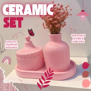 Puede incluir: Un juego de cerámica rosa que incluye un jarrón con flores secas, un tarro con tapa y una bandeja. El jarrón y el tarro tienen un diseño acanalado. El juego está diseñado para flores secas. El texto de la imagen dice "CERAMIC SET".