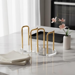 May include: Two clear acrylic and gold metal napkin holders with a minimalist design. The holders have a circular base with vertical gold bars. A folded white napkin and a small vase with pink flowers are also visible.