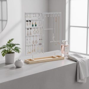 Op de afbeelding: Een witte metalen sieradenorganizer met meerdere haken en gaten voor oorbellen en kettingen. Het heeft een lichte houten basis met twee kleine compartimenten. Een kleine witte keramische ringhouder en een potplant staan in de buurt.