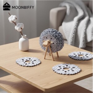 Peut inclure: Une table en bois présente une figurine de mouton décorative avec un motif de flocon de neige gris et trois dessous de verre assortis. Un petit vase blanc contient des tiges de coton. La marque "MOONBIFFY" est visible en haut à gauche.