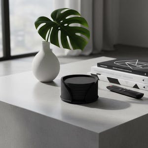 Peut inclure: Scène de salon moderne avec un vase blanc contenant une grande feuille verte, une pile de livres, une télécommande et un ensemble de dessous de verre noirs. Le tout est disposé sur une table basse grise claire.