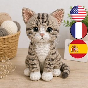 Puede incluir: Una figura de gato de ganchillo hecha a mano en tonos marrón, beige y blanco. El gato tiene ojos grandes y expresivos y marcas rayadas. Las orejas están forradas de rosa. El gato está sentado erguido con la cola enrollada.