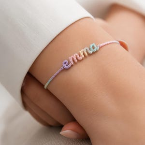 Può includere: Un braccialetto dai colori pastello con il nome "emma" scritto in corsivo. Il braccialetto è realizzato in un materiale intrecciato nelle tonalità del rosa, viola, blu e giallo. Il braccialetto è indossato al polso.