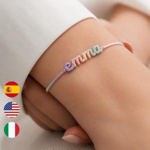 以下が含まれることがあります： パステルカラーのブレスレットで、名前は「emma」です。ブレスレットは、ピンク、紫、黄色、青の色合いの織り糸で作られています。ブレスレットは手首に着用されています。
