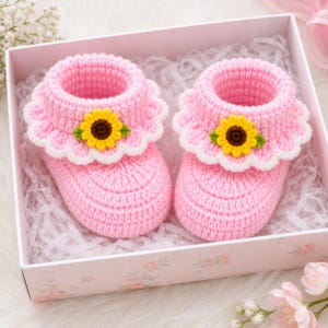 May include: A pair of light pink crochet baby booties featuring a white scalloped cuff and a small yellow sunflower applique with green leaves on the front of each shoe, presented inside a light pink gift box with white shredded paper filler.