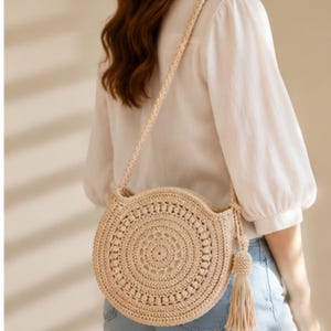 Puede incluir: Un bolso redondo de ganchillo beige con una correa larga y una borla. El bolso tiene un diseño calado detallado y se lleva al hombro. El bolso está hecho de una fibra natural.