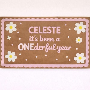 May include: A brown rectangular sign with a pink scalloped border and white daisy accents. The sign reads "CELESTE it's been a ONEderful year" in pink lettering. White dots are scattered around the text and flowers.