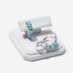 May include: Silver hoop earrings with light blue square gemstones. One earring is displayed on a white jewelry stand, while a pair of earrings rests on the stand's base. The earrings are set against a white background.