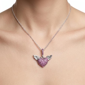 May include: A silver necklace featuring a heart-shaped pendant with pink gemstones and small wings. The pendant hangs from a delicate chain. The heart is filled with small, round pink stones.