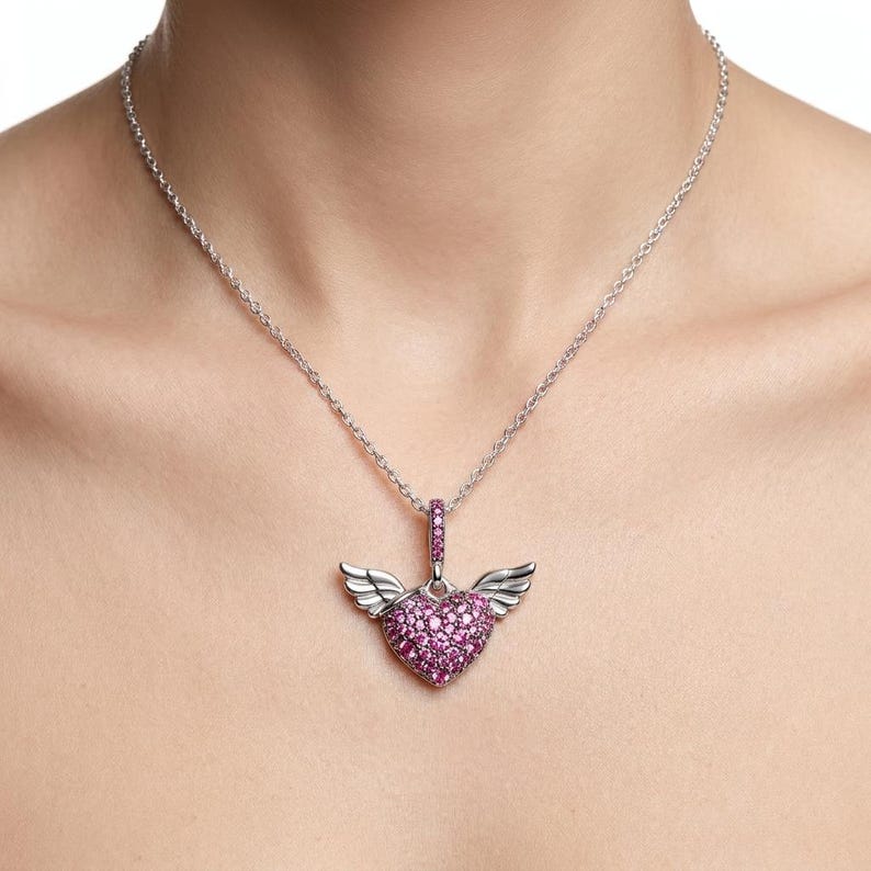 May include: A silver necklace featuring a heart-shaped pendant with pink gemstones and small wings. The pendant hangs from a delicate chain. The heart is filled with small, round pink stones.