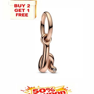 Puede incluir: Charm de color oro rosa con forma de la letra "J" con una pequeña gema transparente. El charm tiene un lazo en la parte superior para sujetarlo. Una caja de regalo con un lazo dorado y el texto "BUY 2 GET 1 FREE" está en el fondo.