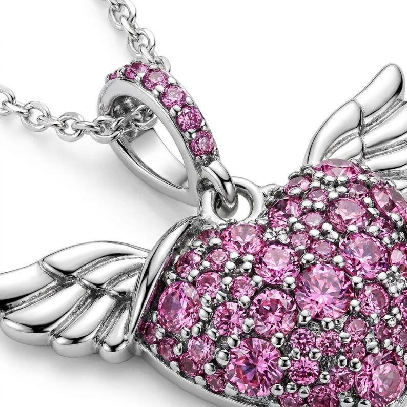 May include: A silver-toned necklace with a heart-shaped pendant. The pendant is encrusted with pink gemstones and features wing-like details. The chain is delicate and the pendant is in focus.