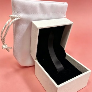 May include: An open white jewelry box with a black velvet interior and a black ribbon. A white drawstring pouch is next to the box. The background is a solid pink color.