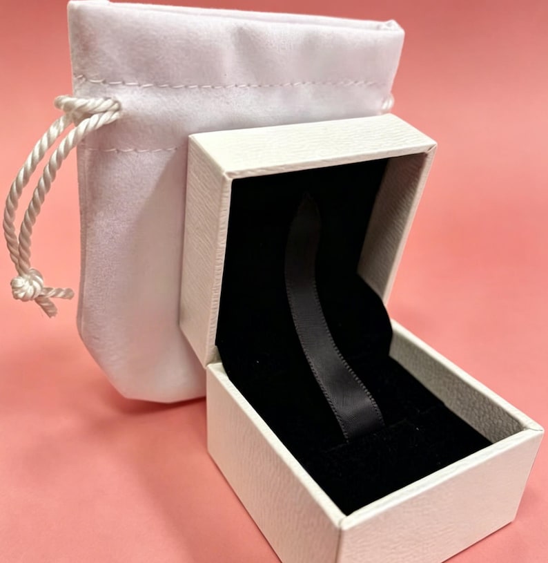 May include: An open white jewelry box with a black velvet interior and a black ribbon. A white drawstring pouch is next to the box. The background is a solid pink color.