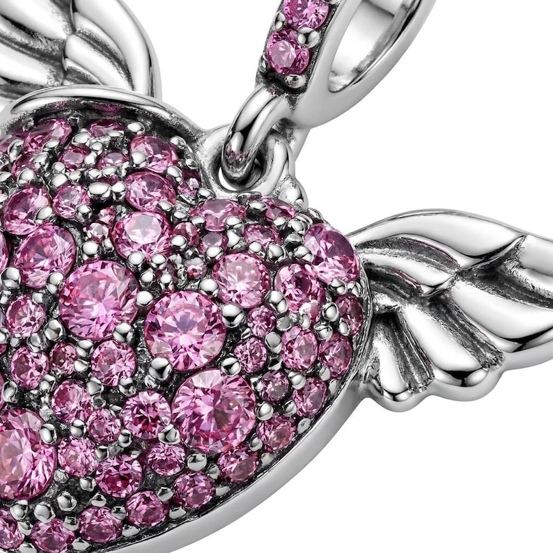 May include: A close-up of a silver charm with a heart shape, covered in pink gemstones. The heart has small, detailed wings on either side. The charm is designed for a bracelet or necklace.