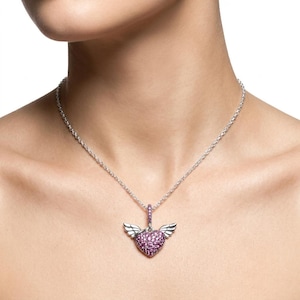 May include: A silver necklace featuring a pendant with a heart-shaped design. The heart is encrusted with small, pink gemstones and has small, silver wings on either side. The necklace is worn around the neck.