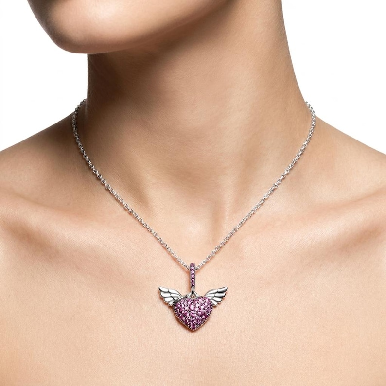 May include: A silver necklace featuring a pendant with a heart-shaped design. The heart is encrusted with small, pink gemstones and has small, silver wings on either side. The necklace is worn around the neck.