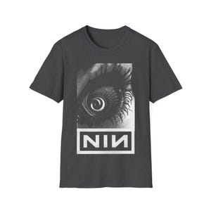May include: Dark grey t-shirt with a black and white eye graphic, spiral iris and long eyelashes. The white text "NIN" is in a rectangular box below the eye. The shirt has a crew neck.