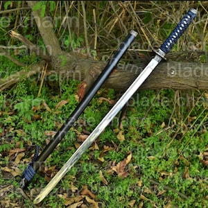 May include: A katana sword with a silver blade and a black scabbard. The handle is wrapped in blue cord. The sword is lying on a bed of green foliage and a fallen tree branch.