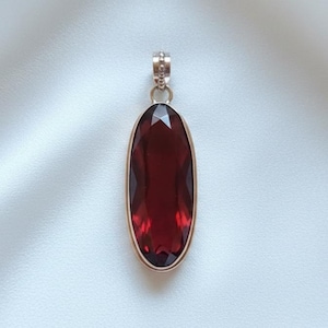 May include: An oval-shaped pendant featuring a large, faceted, deep red gemstone. The gemstone is set in a gold-coloured metal frame and has a small, decorative bail at the top. The pendant is shown against a white background.