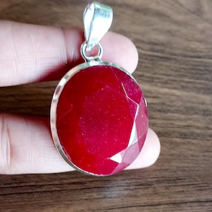 May include: A round, faceted ruby pendant with a silver-coloured bail. The deep red gemstone is set in a silver bezel. The pendant is held in a hand against a blurred wooden background. A striking piece of jewellery.