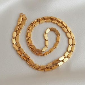 May include: A gold-coloured necklace with a textured, rectangular link design. The necklace is arranged in a spiral pattern on a white, satin-like surface. The clasp is visible at the top of the image.