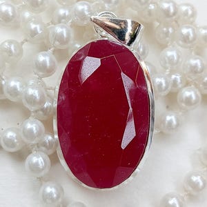 May include: A silver pendant featuring a large, faceted, oval-cut, deep red gemstone. The pendant is set against a backdrop of white pearls, contrasting the shiny metal, the red stone, and the pearls' lustre.