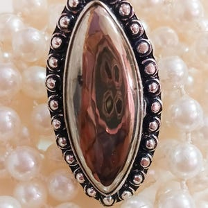 Puede incluir: Un anillo en forma de marquesa con una piedra central metálica lisa y reflectante, engastada en un marco de plata oxidada oscura decorado con un borde de cuentas, sobre un conjunto de perlas blancas redondas y brillantes.