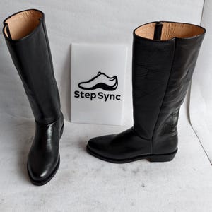 May include: Two black leather knee-high boots with a pointed toe and a low heel. The boots have a smooth, shiny finish and a light-colored interior. A white sign with the text "Step Sync" and a shoe graphic is between the boots.