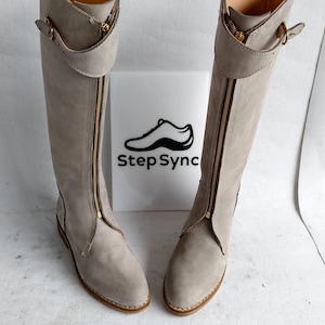 May include: A pair of light gray knee-high boots with a zipper down the front. The boots have a gold-colored zipper and buckle details. The boots are displayed in front of a white sign with the text "Step Sync" and a shoe graphic.
