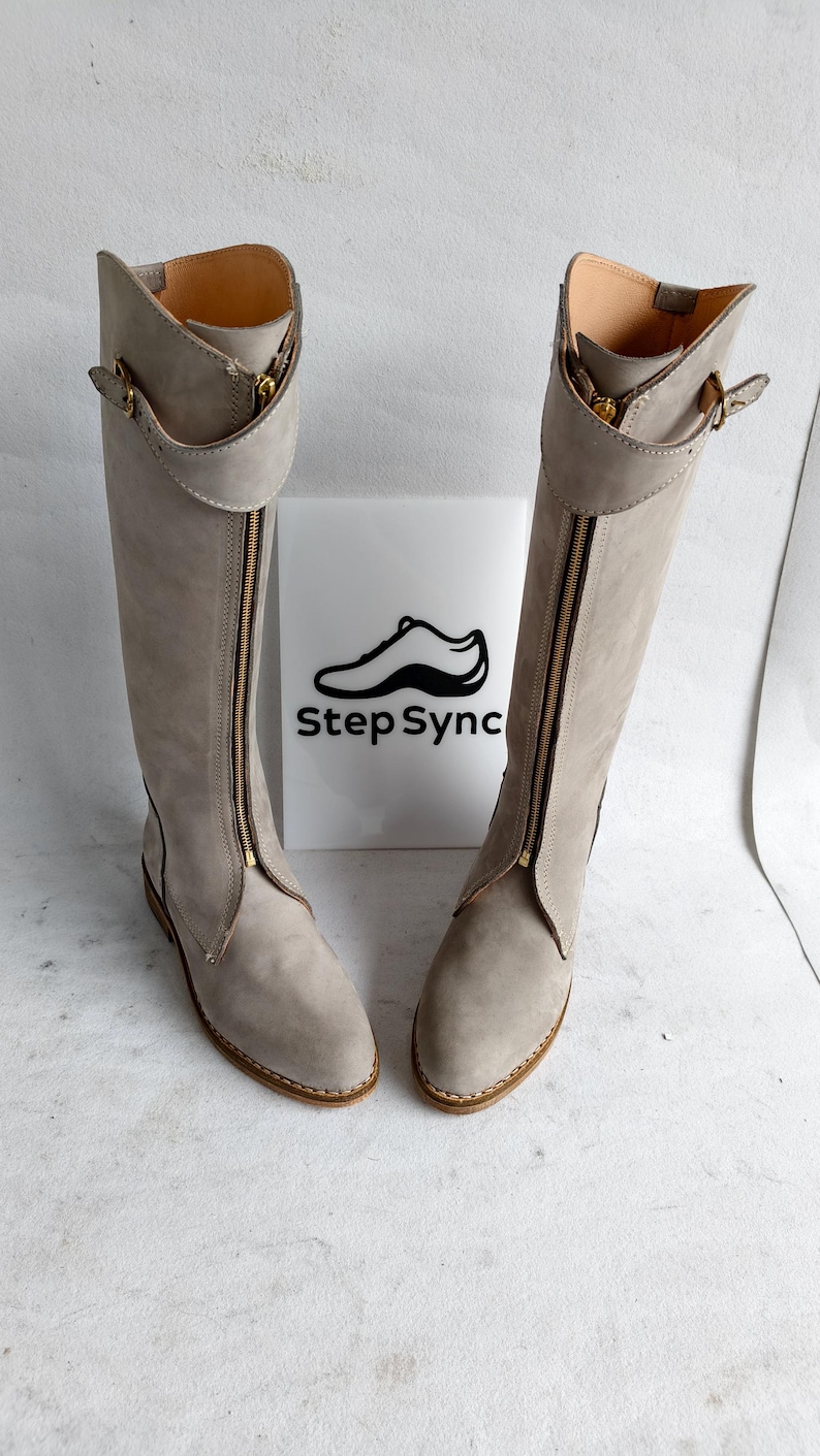 May include: A pair of light gray knee-high boots with a zipper down the front. The boots have a gold-colored zipper and buckle details. The boots are displayed in front of a white sign with the text "Step Sync" and a shoe graphic.