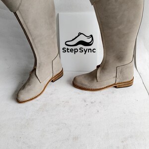 May include: A pair of tall, light gray riding boots with a side zipper and buckle detail. The boots have a flat sole and are made of a suede-like material. The logo "Step Sync" is visible on a white background.