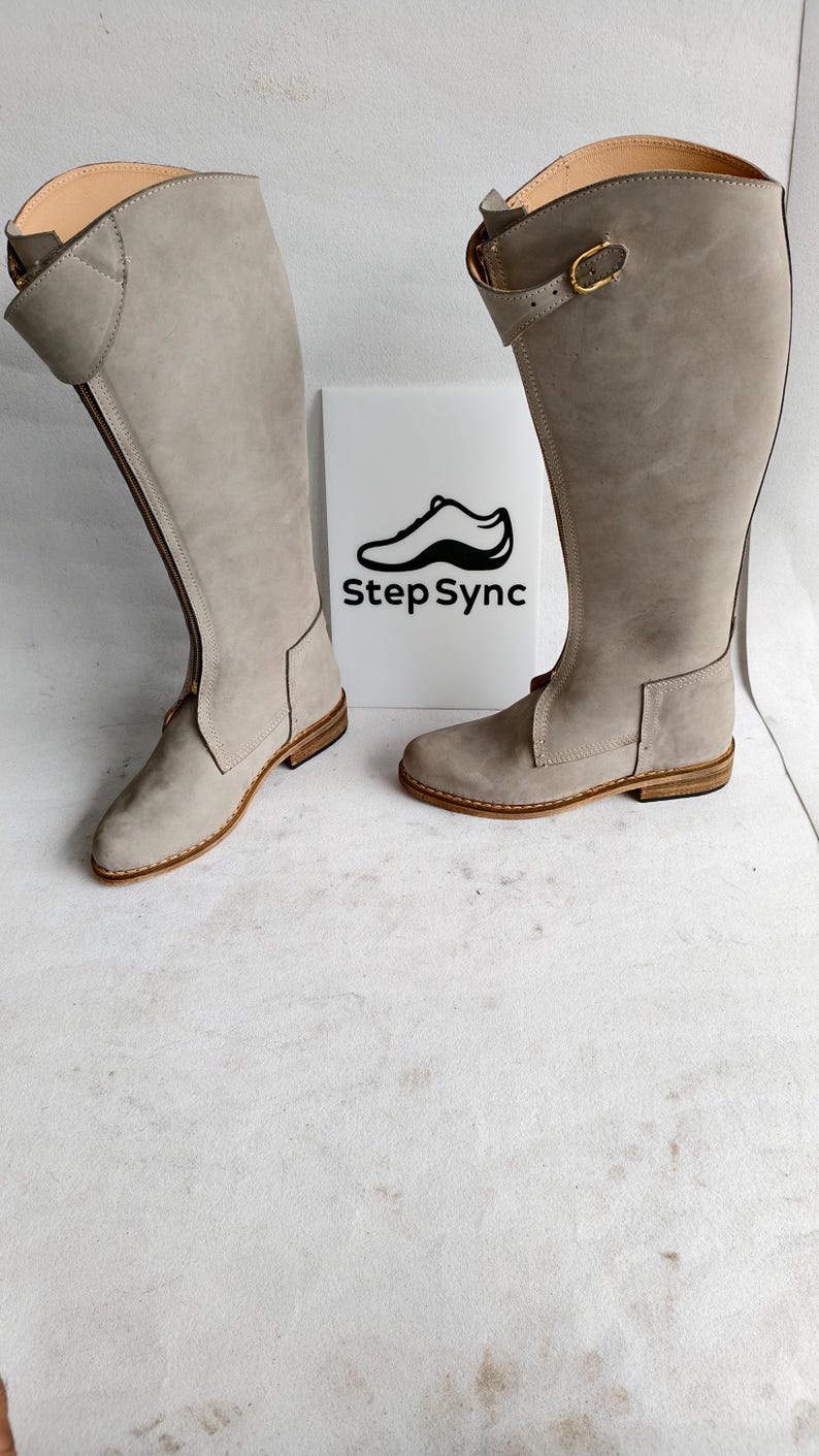 May include: A pair of tall, light gray riding boots with a side zipper and buckle detail. The boots have a flat sole and are made of a suede-like material. The logo "Step Sync" is visible on a white background.