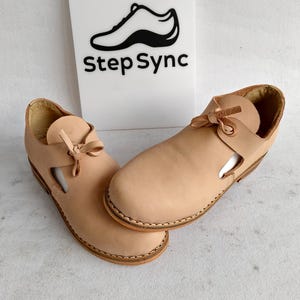 Op de afbeelding: Een paar lichtbruine leren sandalen met een uitgesneden ontwerp en strikdetail. De sandalen worden gepresenteerd op een licht oppervlak, met een wit bord op de achtergrond met de tekst "Step Sync" en een schoenenlogo.