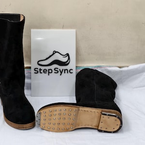 Puede incluir: Dos botas de ante negro, una alta y otra a la altura del tobillo, se exhiben junto a un letrero con el texto "Step Sync" y un gráfico de zapato. Las botas están sobre una superficie blanca.