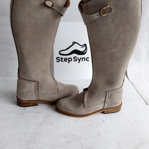 May include: A pair of tall, gray leather boots with a buckle detail. The boots have a knee-high design and a light brown sole. The boots are displayed in front of a white sign with the text "Step Sync" and a shoe logo.