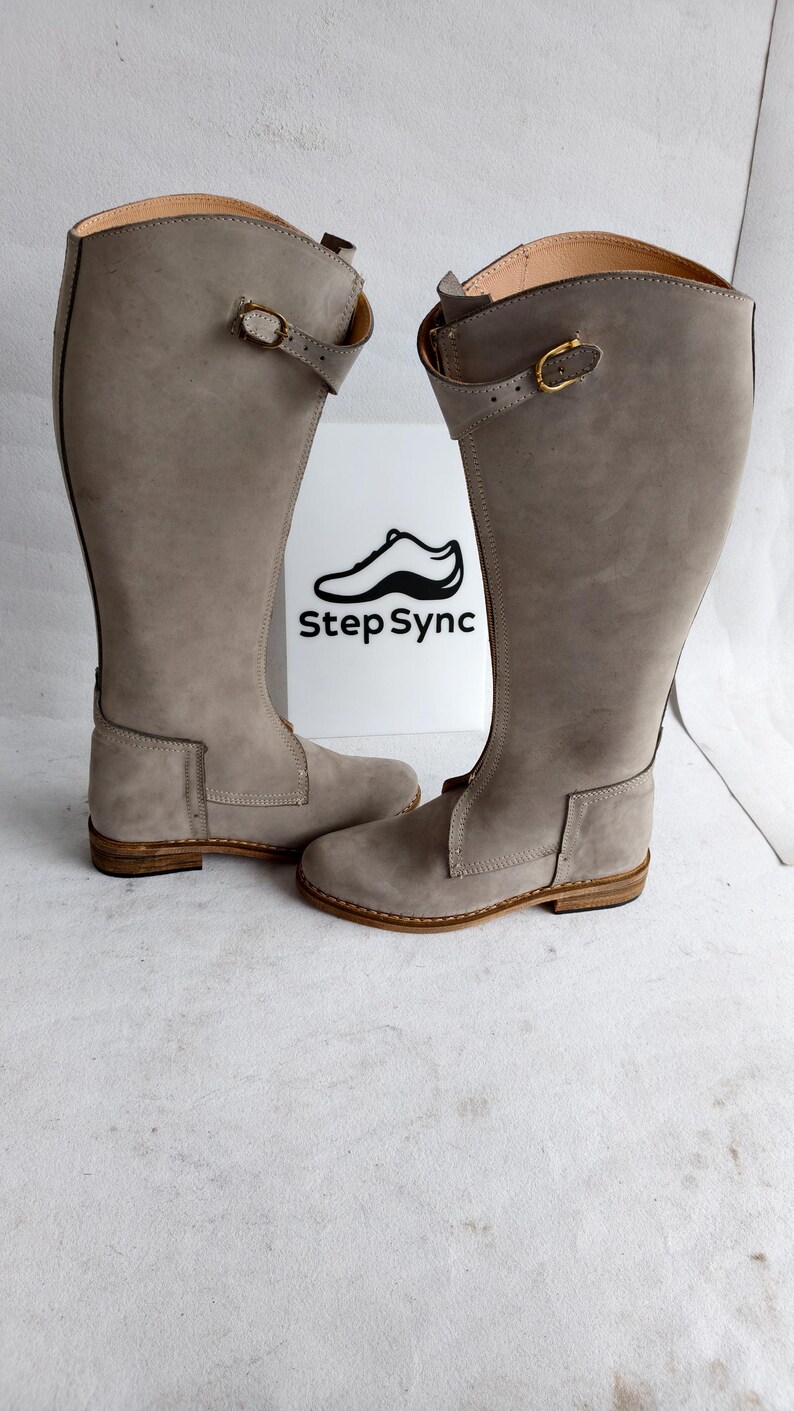May include: A pair of tall, gray leather boots with a buckle detail. The boots have a knee-high design and a light brown sole. The boots are displayed in front of a white sign with the text "Step Sync" and a shoe logo.