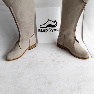May include: A pair of light gray suede knee-high boots with a zipper closure. The boots have a rounded toe and a small heel. The brand name "Step Sync" is printed on a white sign between the boots.