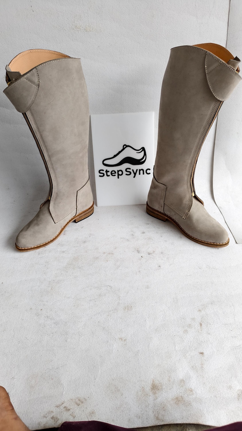May include: A pair of light gray suede knee-high boots with a zipper closure. The boots have a rounded toe and a small heel. The brand name "Step Sync" is printed on a white sign between the boots.