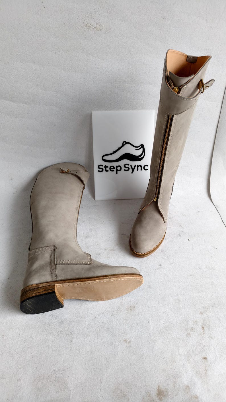 May include: Two gray leather riding boots with a side zipper and buckle detail. The boots are displayed on a white surface with a sign that reads "Step Sync" and a shoe logo.