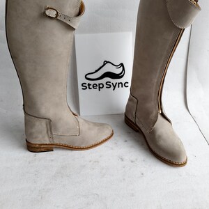 May include: A pair of tall, light gray boots with a side zipper and buckle detail. The boots are made of a suede-like material and have a low heel. The boots are displayed in front of a white sign with the text "Step Sync".