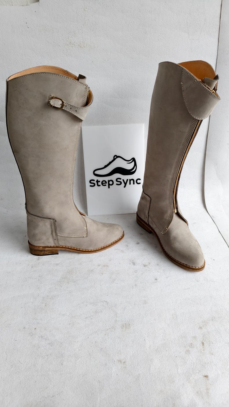 May include: A pair of tall, light gray boots with a side zipper and buckle detail. The boots are made of a suede-like material and have a low heel. The boots are displayed in front of a white sign with the text "Step Sync".