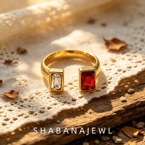 Pode incluir: Um anel aberto em tom dourado com duas pedras retangulares, uma transparente e outra vermelho-escura, fixadas em cada extremidade da banda. O texto SHABANAJEWL está impresso na parte inferior da imagem. O anel tem um acabamento polido e design.