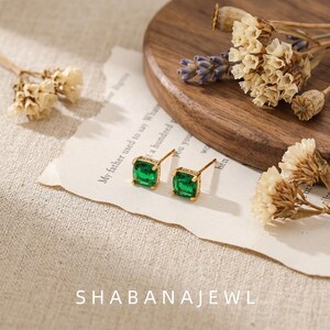May include: A pair of gold stud earrings featuring square-cut green gemstones. The earrings are positioned on a piece of torn paper with printed text, accompanied by dried flowers. The text SHABANAJEWL is visible at the bottom of the image.