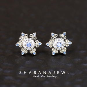 May include: A pair of silver snowflake stud earrings with blue and clear gemstones. The earrings have a detailed, ornate design with a central blue stone and smaller blue accents. The text "SHABANAJEWL Handcrafted Jewellery" is at the bottom.