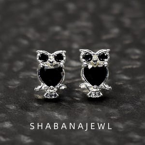Puede incluir: Un par de pendientes de botón con forma de búho, con dos pequeñas piedras negras circulares para los ojos y una piedra negra más grande en forma de corazón para el cuerpo. El texto SHABANAJEWL aparece en blanco en la parte inferior.