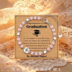 May include: A beaded bracelet featuring light pink round beads and silver-toned square spacers, with a circular charm displaying a compass design. The bracelet is displayed on a card with the text Congratulations on your Graduation.